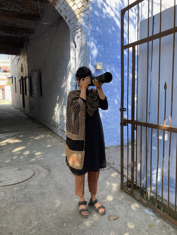 India Street Lettering founder, Pooja Saxena, is standing in an alley holding her camera up, which is pointing to a painted sign that is not in the frame.