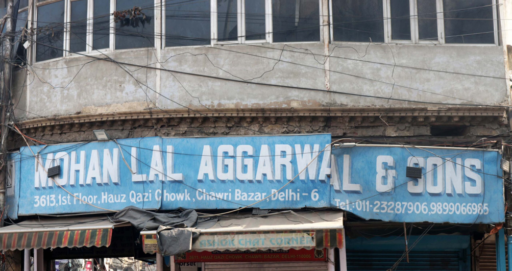 On a grey, rundown building there is a blue painted sign that reads Mohan Lal Aggarwal & Sons. The letters are painted in uppercase and in white. They have a two-tone close shade.